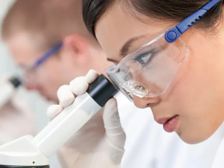 A researcher looks into a microscope. She is wearing protective goggles.