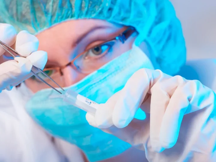 A researcher holds tweezers above a plastic vial. She is wearing protective goggles and a face mask.