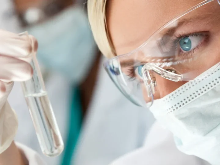 A researcher examines a test tube of clear liquid. She is wearing protective goggles and a face mask.